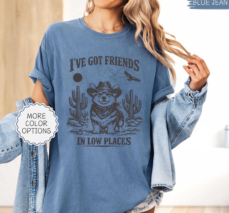 May include: Blue jean-colored t-shirt with a graphic featuring a Corgi wearing a cowboy hat, cacti, and the text "I've Got Friends In Low Places." The shirt has rolled-up sleeves and a relaxed fit.