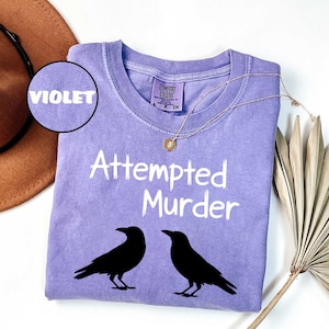 May include: A violet-colored Comfort Colors t-shirt with the words "Attempted Murder" in white, along with two black raven silhouettes. A gold necklace and a brown hat are also visible.