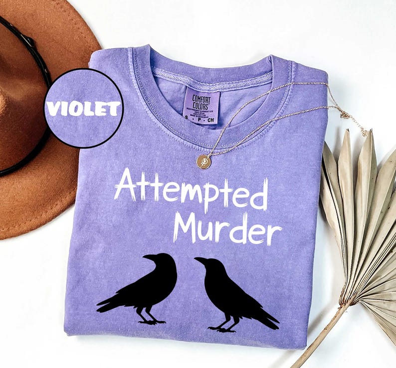 May include: A violet-colored Comfort Colors t-shirt with the words "Attempted Murder" in white, along with two black raven silhouettes. A gold necklace and a brown hat are also visible.