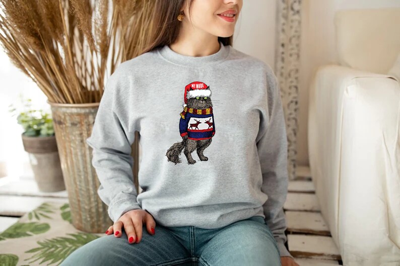 K&ouml;nnte beinhalten: Ein graues Sweatshirt mit einem Aufdruck eines Katers, der einen Weihnachtsmann-Hut und einen Weihnachtspullover mit Rentieren tr&auml;gt. Der Text auf dem Hut lautet "My Mama".