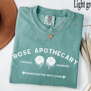 May include: Light green t-shirt with the text "ROSE APOTHECARY" and two rose illustrations. The shirt also has the words "LOCALLY SOURCED HANDCRAFTED WITH CARE". A silver necklace with a pendant is on the shirt.
