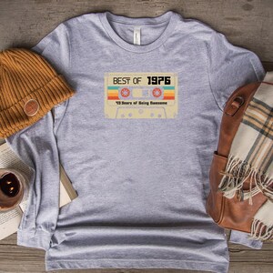 May include: A heather grey long-sleeved t-shirt with a vintage cassette tape design. The tape has the text "Best of 1976" and "49 Years of Being Awesome".
