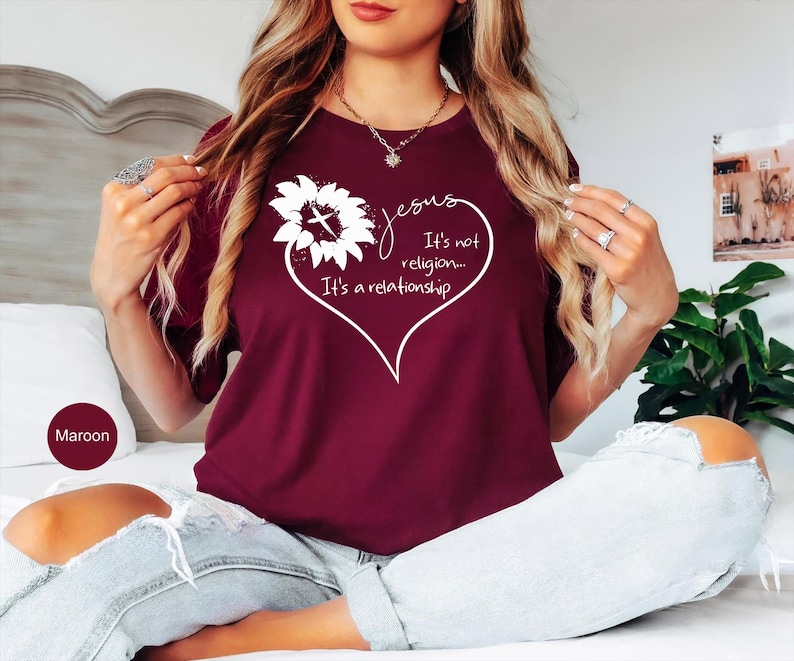 May include: A maroon t-shirt featuring a white graphic of a sunflower with a cross in the center, shaped into a heart outline. The text reads: Jesus It's not religion... It's a relationship. The shirt has short sleeves and a crew neckline.