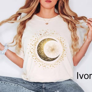 May include: Ivory-colored t-shirt featuring a gold crescent moon and star design. The moon has an ornate, swirling pattern and a bright center. The design is surrounded by smaller gold stars.