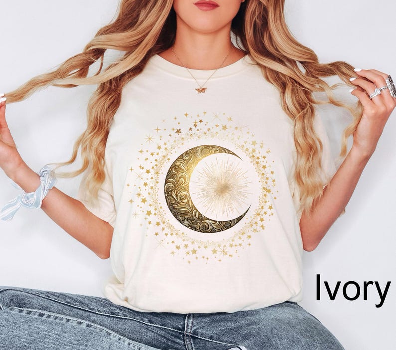 May include: Ivory-colored t-shirt featuring a gold crescent moon and star design. The moon has an ornate, swirling pattern and a bright center. The design is surrounded by smaller gold stars.