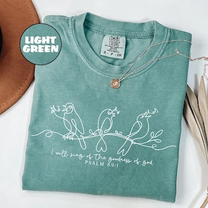 May include: Light green t-shirt featuring a white line drawing of three birds perched on a branch. The text "I will sing of the goodness of god, PSALM 89:1" is printed below. A silver necklace with a pendant rests on the shirt.
