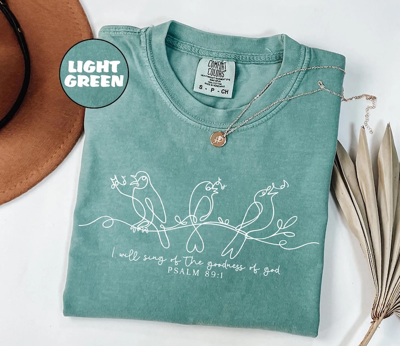 May include: Light green t-shirt featuring a white line drawing of three birds perched on a branch. The text "I will sing of the goodness of god, PSALM 89:1" is printed below. A silver necklace with a pendant rests on the shirt.