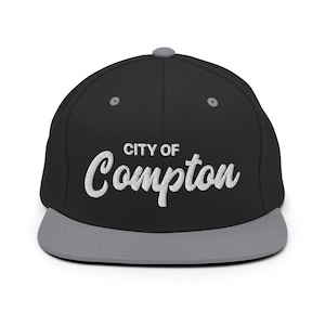 May include: Black and gray baseball cap with the text "CITY OF Compton" embroidered in white on the front.