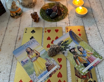 May include: A tarot card reading spread with four cards laid out on a white surface. The cards are gold with images of people and symbols. There are two lit candles and a small snow globe with an angel figurine on the surface.