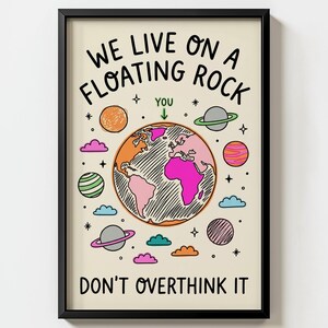 Könnte beinhalten: Ein gerahmter Kunstdruck auf cremefarbenem Hintergrund mit einer zentralen Erddarstellung mit rosa und orangefarbenen Kontinenten. Der Text lautet WE LIVE ON A FLOATING ROCK, YOU und DON'T OVERTHINK IT. Um die Erde herum sind bunte Planeten, Sterne und Wolken abgebildet.