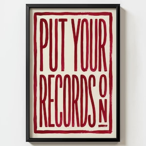 Op de afbeelding: Een ingelijste kunstdruk met de tekst "PUT YOUR RECORDS ON" in vetgedrukte, rode letters. De tekst staat centraal op een crèmekleurige achtergrond, omlijst door een rode rand en een zwart frame. Het kunstwerk heeft een vintage uitstraling.
