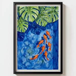 May include: Watercolor painting of orange koi fish swimming in blue water under green leaves. The artwork is framed in a black frame. The painting has a tropical theme and is a decorative piece.
