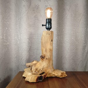 Puede incluir: Una l&aacute;mpara de mesa de madera con una base de madera natural y una bombilla. La base tiene forma de tronco de &aacute;rbol con ramas.