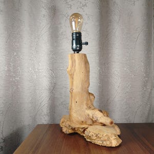 Puede incluir: Una l&aacute;mpara de mesa de madera r&uacute;stica con una base de madera natural y un casquillo de luz negro. La l&aacute;mpara est&aacute; iluminada con una bombilla transparente.