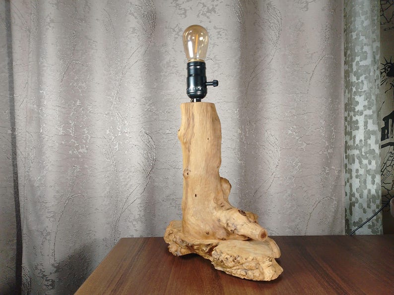 Puede incluir: Una l&aacute;mpara de mesa de madera r&uacute;stica con una base de madera natural y un casquillo de luz negro. La l&aacute;mpara est&aacute; iluminada con una bombilla transparente.