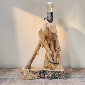 May include: A rustic wooden table lamp with a natural, organic design. The lamp features a light bulb in a metal socket atop a carved wooden base. The base is made from a tree trunk with branches extending out. The lamp is approximately 46 cm tall.