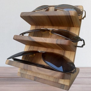 May include: A three-tiered wooden display stand featuring a natural grain pattern, designed to hold eyewear. The stand is angled to support three pairs of sunglasses, each resting on its own horizontal wooden shelf with a smooth, polished finish.