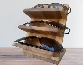May include: A three-tiered wooden display stand featuring a natural grain pattern, designed to hold eyewear. The stand is angled to support three pairs of sunglasses, each resting on its own horizontal wooden shelf with a smooth, polished finish.