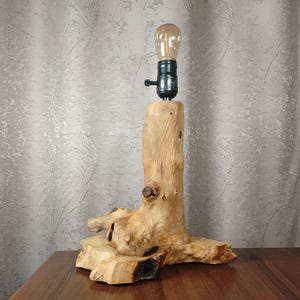 Puede incluir: Una l&aacute;mpara de mesa de madera r&uacute;stica con una base de madera natural y una pantalla de metal negro. La pantalla tiene una bombilla transparente en el interior.