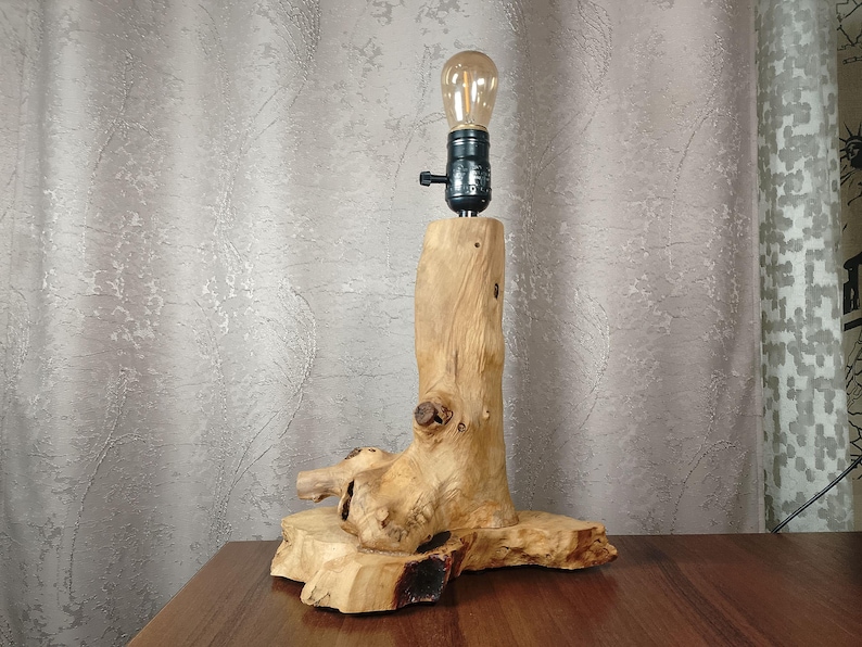 Puede incluir: Una l&aacute;mpara de mesa de madera r&uacute;stica con una base de madera natural y una pantalla de metal negro. La pantalla tiene una bombilla transparente en el interior.