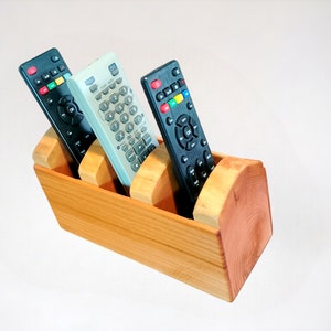 May include: A wooden remote control organizer with three slots. The organizer is made of light brown wood and has a natural finish. Three remotes are in the slots, two black and one light gray.