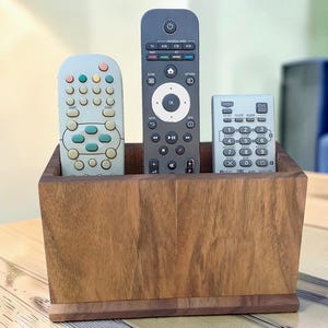 May include: A rectangular wooden remote control holder with a natural brown wood grain finish. The holder contains three remote controls: a light grey remote on the left, a tall dark grey remote in the centre, and a small light grey remote on the right.