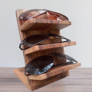 Op de afbeelding: Een houten displaystandaard met drie niveaus en horizontale planken met een natuurlijke houtnerfafwerking. Drie zonnebrillen met donkere glazen rusten op de planken, met één paar op elk niveau van de schuin geplaatste structuur.