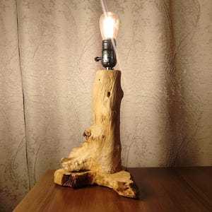 Puede incluir: Una l&aacute;mpara de mesa de madera con una base de madera natural y una bombilla. La l&aacute;mpara est&aacute; encendida y proyecta un brillo c&aacute;lido.