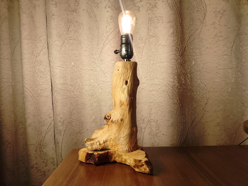 Puede incluir: Una l&aacute;mpara de mesa de madera con una base de madera natural y una bombilla. La l&aacute;mpara est&aacute; encendida y proyecta un brillo c&aacute;lido.