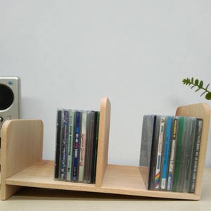 Op de afbeelding: Een lichtgekleurde houten cd-plank met twee compartimenten, elk met verschillende cd's. De plank staat op een lichtgekleurd oppervlak, met een radio en een kleine plant op de achtergrond.