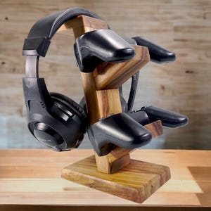 Op de afbeelding: Een houten standaard met een headset en twee controllers. De standaard is gemaakt van lichtbruin hout en heeft een natuurlijke afwerking. De headset is zwart en heeft een microfoon. De controllers zijn zwart en hebben knoppen en joysticks.