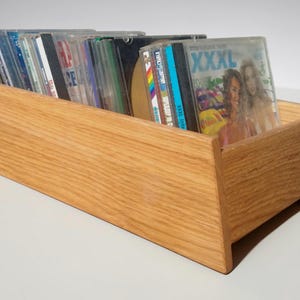 Op de afbeelding: Een houten cd-houder gevuld met diverse cd's. De houder is gemaakt van lichtgekleurd hout en heeft een rechthoekige vorm met schuine zijkanten. De cd's zitten in doorzichtige plastic hoesjes.