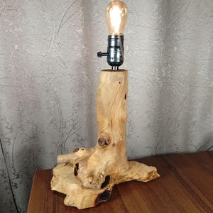 Puede incluir: Una l&aacute;mpara de mesa de madera con una base de madera natural y una bombilla de vidrio transparente. La base tiene forma de tronco de &aacute;rbol con nudos y ramas.