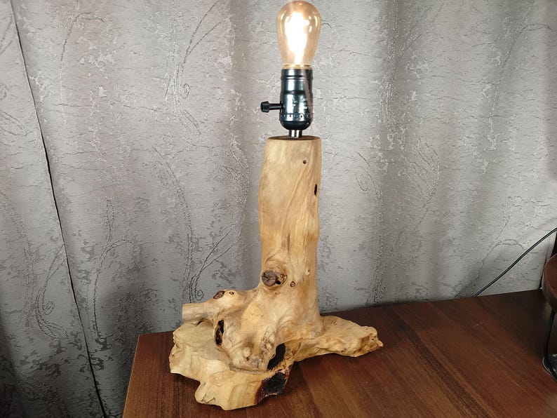 Puede incluir: Una l&aacute;mpara de mesa de madera con una base de madera natural y una bombilla de vidrio transparente. La base tiene forma de tronco de &aacute;rbol con nudos y ramas.