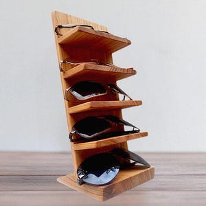 Op de afbeelding: Houten displaystandaard voor zonnebrillen met vijf planken. De standaard is gemaakt van lichtbruin hout en biedt plaats aan meerdere zonnebrillen. De standaard is schuin en heeft een modern design.