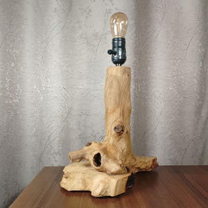 Puede incluir: Una l&aacute;mpara de mesa de madera r&uacute;stica con una base de madera natural y una sencilla luminaria negra. La l&aacute;mpara est&aacute; iluminada con una bombilla transparente.