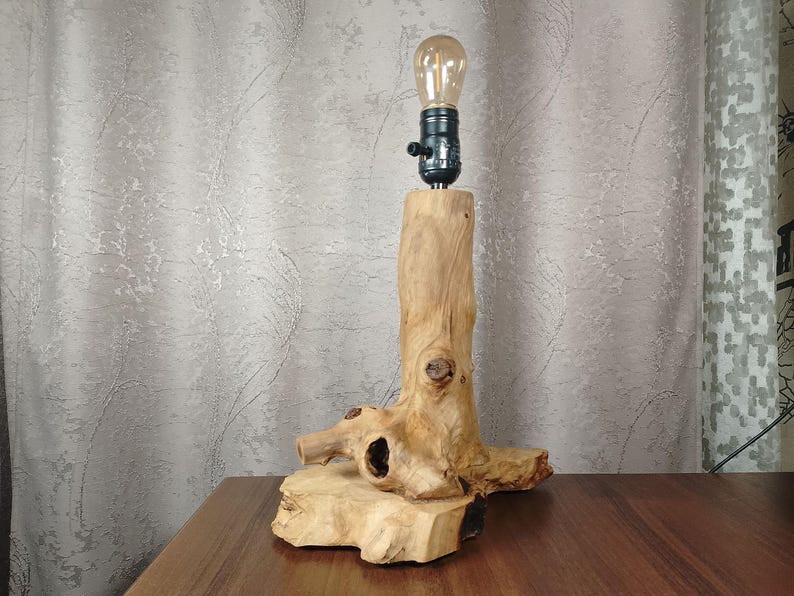 Puede incluir: Una l&aacute;mpara de mesa de madera r&uacute;stica con una base de madera natural y una sencilla luminaria negra. La l&aacute;mpara est&aacute; iluminada con una bombilla transparente.