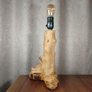 Puede incluir: Una l&aacute;mpara de mesa de madera con una base de madera natural con forma de tronco de &aacute;rbol. La l&aacute;mpara tiene una bombilla de vidrio transparente con una luz blanca c&aacute;lida.