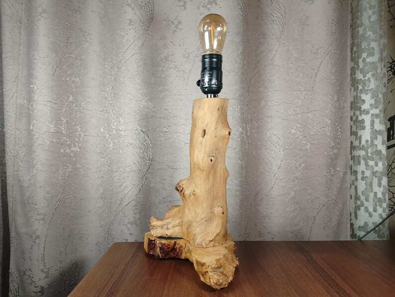 Puede incluir: Una l&aacute;mpara de mesa de madera con una base de madera natural con forma de tronco de &aacute;rbol. La l&aacute;mpara tiene una bombilla de vidrio transparente con una luz blanca c&aacute;lida.