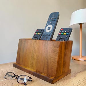 May include: A rectangular wooden remote control holder with a natural grain finish, containing three black remote controls with colorful buttons. A pair of black-rimmed eyeglasses rests on the surface in front of the wooden organizer.