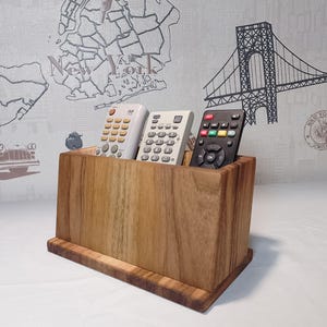 Op de afbeelding: Een houten afstandsbediening organizer met vier vakken voor afstandsbedieningen. De organizer is gemaakt van donkerbruin hout met een natuurlijke houtnerf. Er zitten drie afstandsbedieningen in de organizer, twee witte en een zwarte.