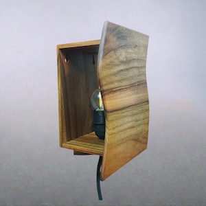 Op de afbeelding: Houten wandlamp met een warme, natuurlijke afwerking. De armatuur heeft een rechthoekig doosontwerp met een gebogen voorpaneel. Een zichtbare gloeilamp is zichtbaar aan de binnenkant en een zwart snoer loopt vanaf de basis.