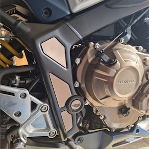 May include: Close-up of a motorcycle engine with a bronze-colored casing and a black and copper-colored protective plate. The plate has a raised logo that says "HONDA".