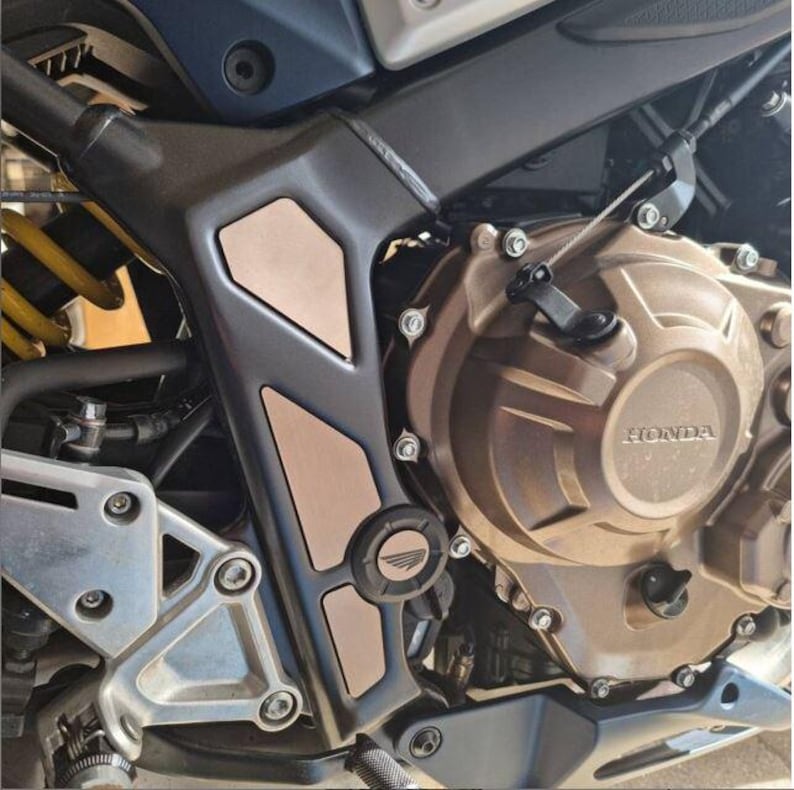May include: Close-up of a motorcycle engine with a bronze-colored casing and a black and copper-colored protective plate. The plate has a raised logo that says "HONDA".