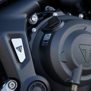 Könnte beinhalten: Ein mattschwarzes Motorradmotorgehäuse mit dem eingeprägten Triumph-Logo auf dem runden Deckel und einem kleinen silbernen dreieckigen Emblem mit demselben Logo an der Seite. Mehrere Metallschrauben halten die dunklen Komponenten zusammen.