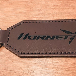 Op de afbeelding: Een bruin leren sleutelhanger met het woord "Hornet" en een gestileerd insectenlogo in zwart. De sleutelhanger heeft een zilveren sleutelring en is rondom de randen gestikt.