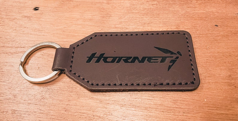 Op de afbeelding: Een bruin leren sleutelhanger met het woord "Hornet" en een gestileerd insectenlogo in zwart. De sleutelhanger heeft een zilveren sleutelring en is rondom de randen gestikt.