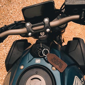 Op de afbeelding: Close-up van een teal motorfiets met een zwart dashboard en stuur. Een bruine leren sleutelhanger met de tekst "Ride Safe" hangt aan het contact. De motorfiets heeft het Honda-logo en de tekst "ENGITECT".