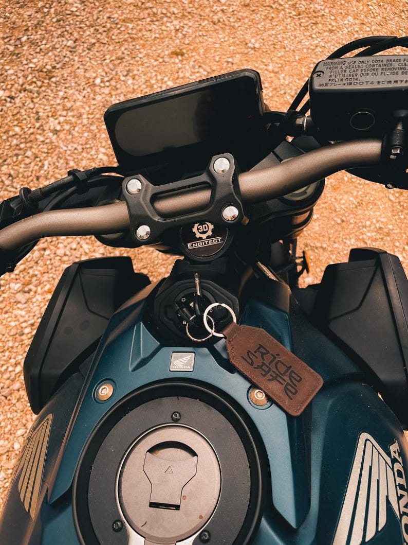 Op de afbeelding: Close-up van een teal motorfiets met een zwart dashboard en stuur. Een bruine leren sleutelhanger met de tekst "Ride Safe" hangt aan het contact. De motorfiets heeft het Honda-logo en de tekst "ENGITECT".