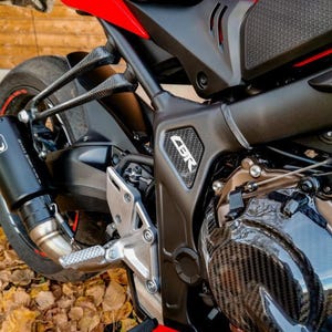 May include: Close-up of a red and black motorcycle with a carbon fiber engine cover. The motorcycle has a black exhaust pipe and a silver footpeg. The word "CBR" is visible on the side of the motorcycle.